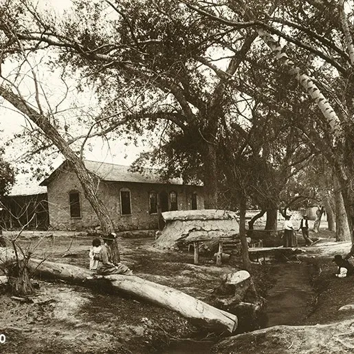 The long adobe ranch house among cottonwood trees, with two people sitting on a fallen log beside the creek